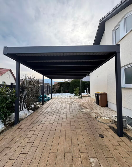 Moderner Carport vor einem Haus mit gepflasterter Einfahrt und trübem Himmel.