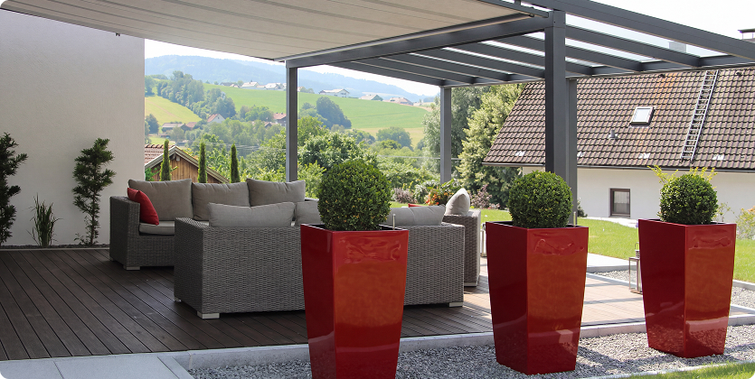 Überdachte Terrasse mit modernen Gartenmöbeln und roten Blumenkübeln vor ländlicher Kulisse.