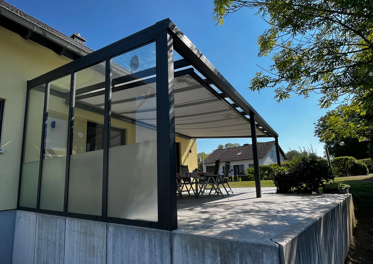 Terrasse mit Glasüberdachung und Gartenmöbeln neben einem Haus in sonniger Umgebung.