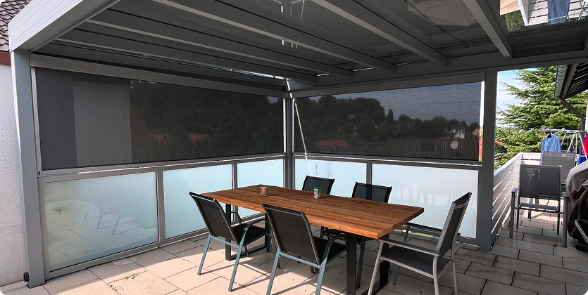 Überdachte Terrasse mit Holztisch, Stühlen und Glaswänden für Sichtschutz.