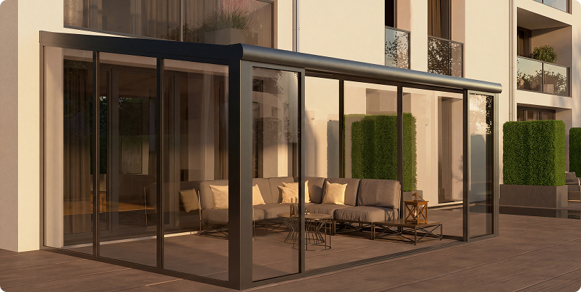 Moderne Terrassenüberdachung mit Glaswänden und Sitzmöbeln vor einem Gebäudekomplex.