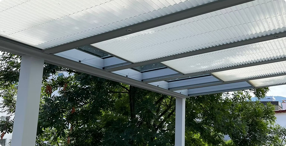 Lichtdurchlässiges Polycarbonatdach über moderner Terrassenkonstruktion mit grünen Bäumen im Hintergrund.