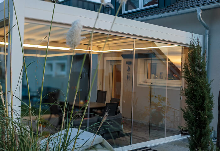 Moderner Wintergarten mit Glaswänden und beleuchteter Terrasse im Freien.
