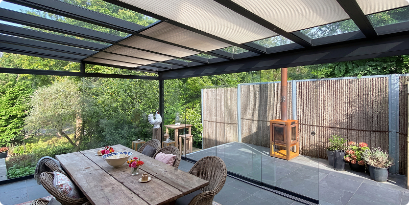 Überdachter Wintergarten mit Holztisch, Rattanstühlen und Glasdach, umgeben von üppiger Vegetation.