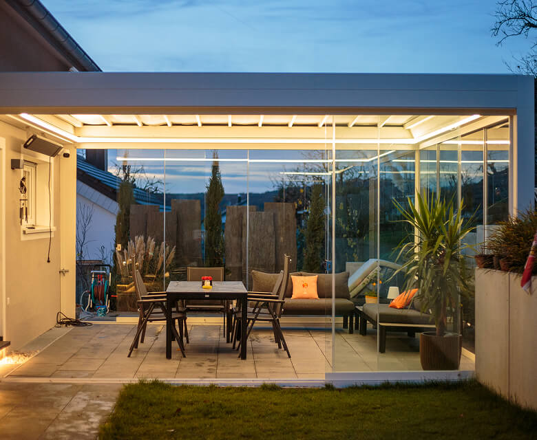 Moderner Wintergarten mit Glaswänden, Möbeln und Beleuchtung bei Abenddämmerung.