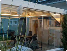 Moderne Terrasse mit Glasüberdachung und Gartenmöbeln, umgeben von Pflanzen.