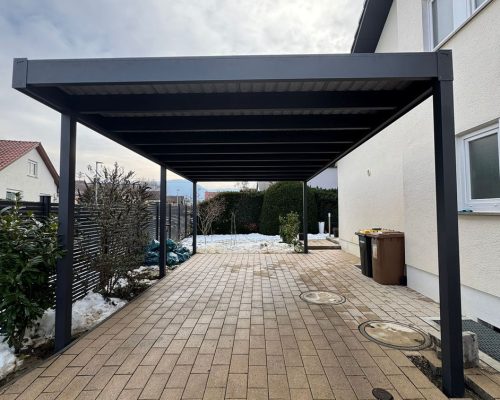 Carport vor einem Haus mit gepflasterter Einfahrt unter bewölktem Himmel.