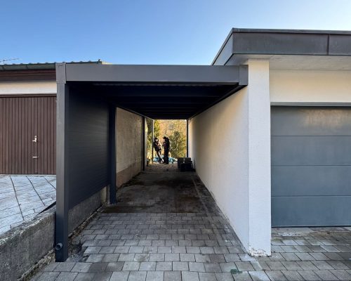 Carport zwischen zwei Gebäuden, mit Pflastersteinen und Personen im Hintergrund.
