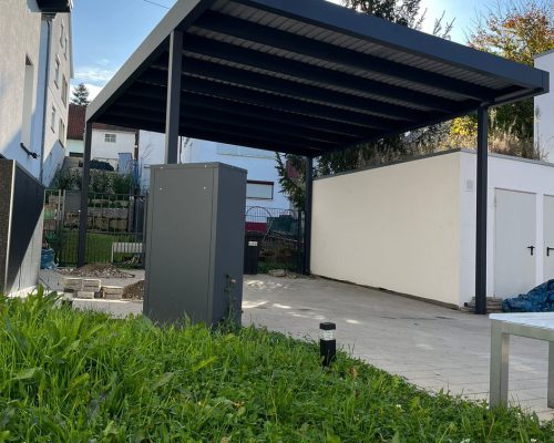 Moderne Carportkonstruktion vor einem Gebäude mit umgebendem Garten und Rasen.