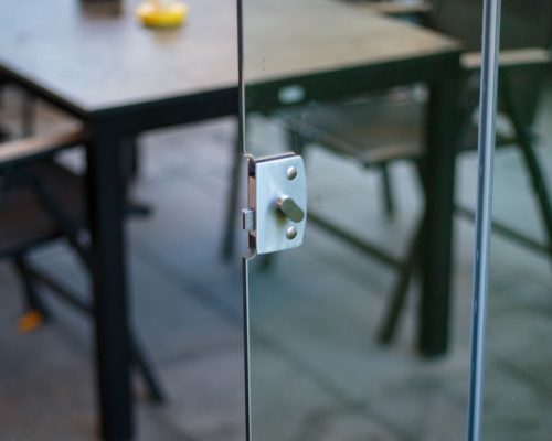Nahaufnahme einer Glastür mit Metallverschluss, im Hintergrund ein Tisch und Stühle.