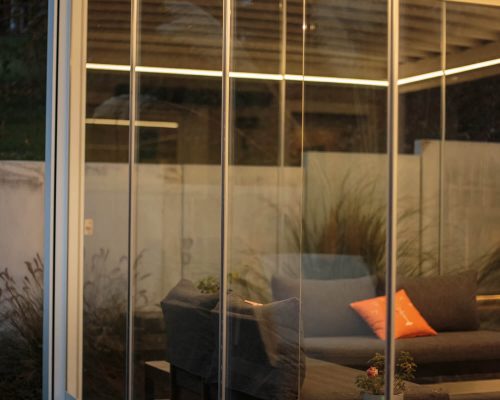 Blick durch Glaswand auf Sofa mit Kissen und Pflanzen im Hintergrund.