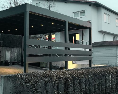 Modernes Haus mit Carport, Terrasse und Hecke im Vordergrund bei Dämmerung.