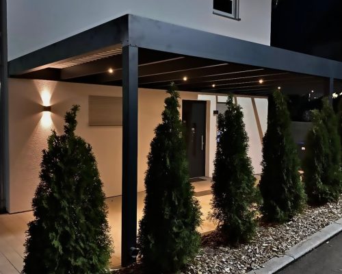 Modernes Haus bei Nacht mit beleuchtetem Eingangsbereich und Pflanzen.