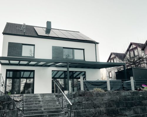 Modernes zweistöckiges Haus mit Solarpanels und Terrasse, umgeben von weiteren Gebäuden.