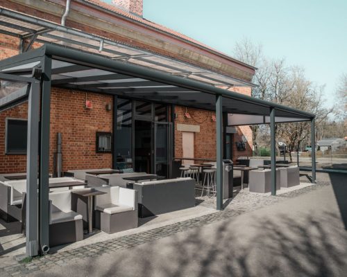 Moderne Terrasse mit Überdachung an einem Backsteingebäude, ausgestattet mit Tischgruppen und Sitzmöglichkeiten.