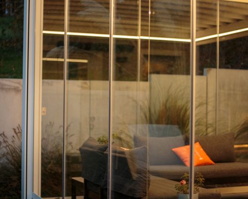 Veranda mit Glaswänden, beleuchtet, Sofa und Pflanzen sichtbar.