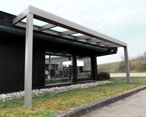 Moderne Glasüberdachung mit Metallstruktur vor einem Gebäude, umgeben von Rasen und Kies.