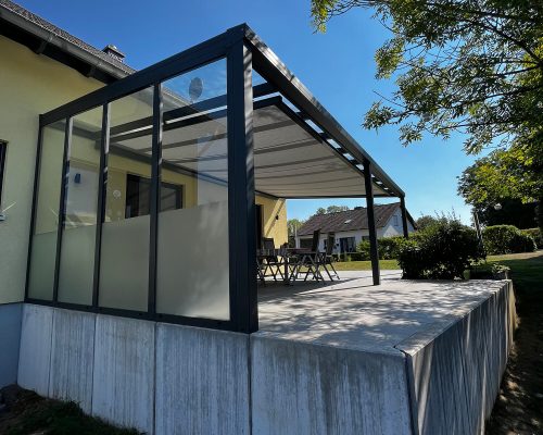 Moderne Terrasse mit Glasüberdachung und Sitzbereich neben einem Wohnhaus, umgeben von Garten.