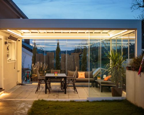 Modern gestaltete Glasveranda mit Gartenmöbeln bei Dämmerung, beleuchtet und umgeben von Pflanzen.