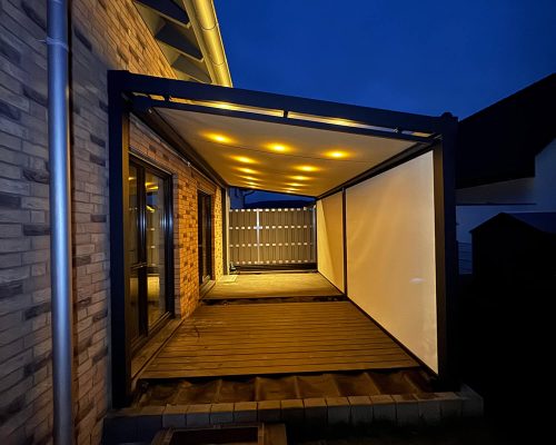 Überdachte Terrasse mit Beleuchtung in der Nacht, an ein Backsteinhaus angrenzend.
