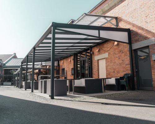 Überdachte Terrasse mit Sitzgelegenheiten vor einem Backsteingebäude bei sonnigem Wetter