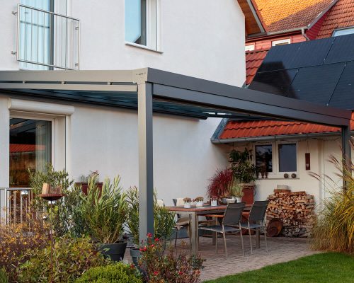 Terrasse mit Möbeln unter Glasüberdachung neben Haus mit Solarpanel und Pflanzen.
