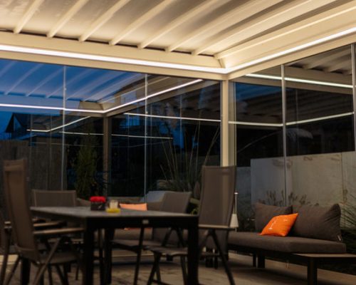 Beleuchtete Terrasse mit Tisch, Stühlen und Sofa, umgeben von Glaswänden bei Nacht.