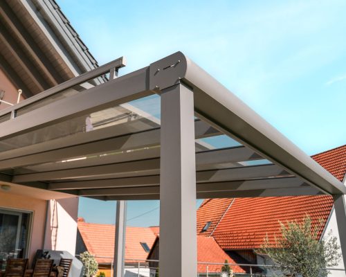 Moderne Terrassenüberdachung mit Glasdach neben Haus mit rotem Dach unter blauem Himmel.