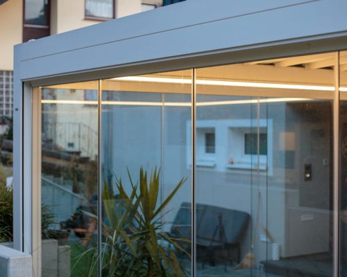 Moderner Wintergarten mit Glaswänden vor einem Haus mit Garten.