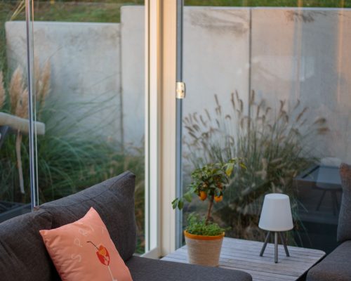 Graues Sofa mit Kissen, Tischlampe und Pflanze vor großer Fensterfront zum Garten.
