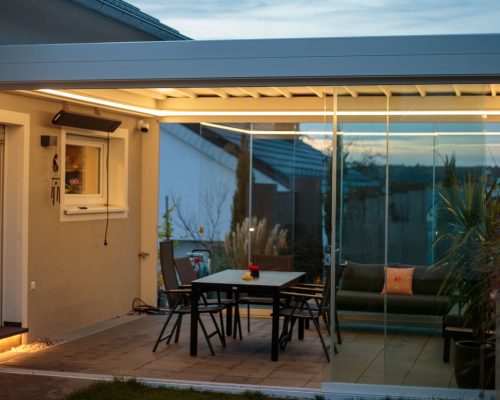 Moderner Wintergarten mit Glaswänden, Tisch und Stühlen, beleuchtet bei Dämmerung.